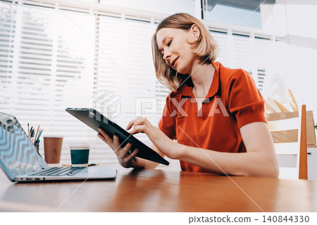 Young woman works on a touchpad, feeling comfortable at cozy home office with a green plant on the background Young woman works on a touchpad, feeling comfortable at cozy home office with a green plant on the background 140844330