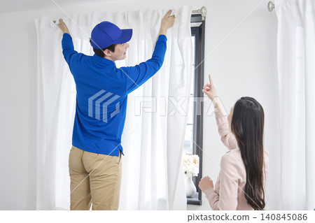 The moving company installs the curtains while a woman gives instructions. The moving company installs the curtains while a woman gives instructions. 140845086