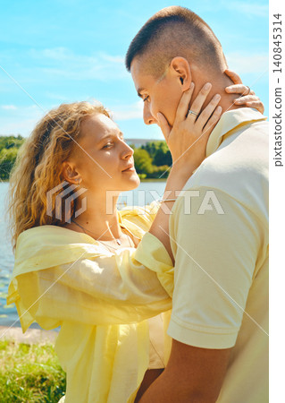 Couple shares moment by lake under blue sky during sunny day near green trees Couple shares moment by lake under blue sky during sunny day near green trees 140845314