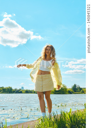Woman stands by lake wearing yellow shirt and skirt on sunny day with clouds in sky Woman stands by lake wearing yellow shirt and skirt on sunny day with clouds in sky 140845321