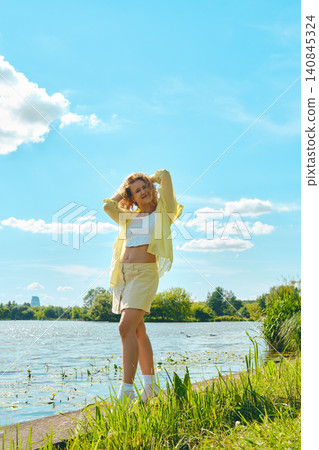 Young woman enjoying sunny day by lake in park with green grass and clouds in blue sky 140845324