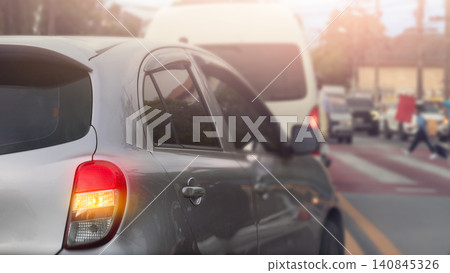 Side rear profile view of a grey car waiting in heavy city traffic during the day. Bright sunlight flares from the upper corner illuminating the busy urban road scene. Blurry of a child crossing road. 140845326