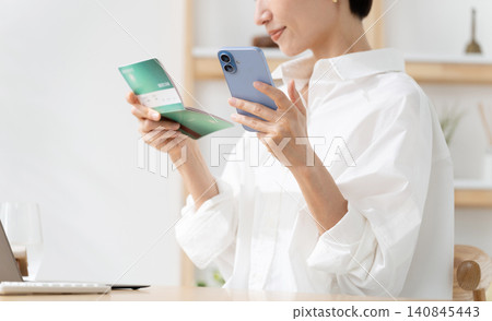 A woman looking at her smartphone and bankbook. Savings, finance, apps, bank, money, household finances, cashless payments, electronic money A woman looking at her smartphone and bankbook. Savings, finance, apps, bank, money, household finances, cashless payments, electronic money 140845443