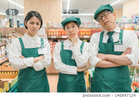 Three store employees stand with their arms crossed, showing their displeasure in the supermarket aisle. Three store employees stand with their arms crossed, showing their displeasure in the supermarket aisle. 140847124