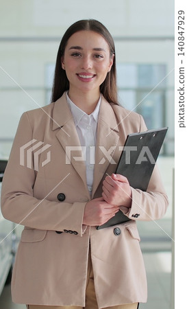 Vertical video Happy woman manager at a car dealership in a business suit holding clipboard documents in her hands stands near the cars. Concept of car shopping, buying, and dealership. Vertical video Happy woman manager at a car dealership in a business suit holding clipboard documents in her hands stands near the cars. Concept of car shopping, buying, and dealership. 140847929