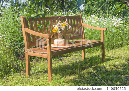 Straw hat and bouquet of whtie daisy flowers in the straw bag on a wooden bench on a blooming meadow in the summer garden by sunny day Straw hat and bouquet of whtie daisy flowers in the straw bag on a wooden bench on a blooming meadow in the summer garden by sunny day 140850323