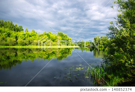 Serene river reflecting lush green trees under a dramatic cloudy sky 140852374
