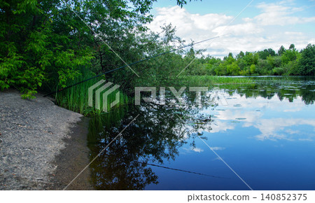 Fishing rod cast into a calm lake surrounded by lush green trees and vegetation 140852375