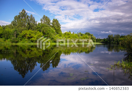 Serene river reflecting lush green trees and cloudy sky on a bright summer day Serene river reflecting lush green trees and cloudy sky on a bright summer day 140852383