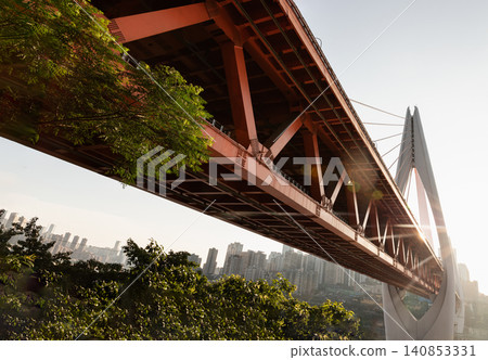 A stunning juxtaposition of nature and modern mega-infrastructure. Lush green tree foliage frames the bold geometric steel structure of the Dongshuimen / Qiansimen Bridge system, with the dense A stunning juxtaposition of nature and modern mega-infrastructure. Lush green tree foliage frames the bold geometric steel structure of the Dongshuimen / Qiansimen Bridge system, with the dense 140853331