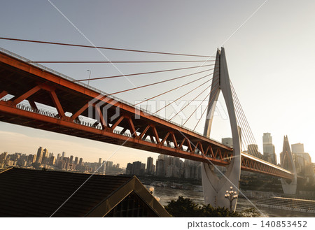 the bold geometry of the Qiansimen Bridge dominates the composition; its vivid orange-red truss structure and towering white pylon sweep across the frame against a hazy, sprawling Chongqing skyline. the bold geometry of the Qiansimen Bridge dominates the composition; its vivid orange-red truss structure and towering white pylon sweep across the frame against a hazy, sprawling Chongqing skyline. 140853452