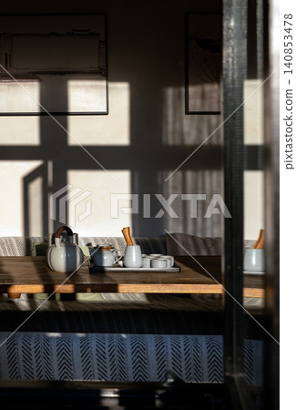 a traditional tea set bathed in sharp, geometric slivers of afternoon sun. The clean lines of the wooden furniture, paired with understated framed artwork and a monochrome patterned bench, evoke a a traditional tea set bathed in sharp, geometric slivers of afternoon sun. The clean lines of the wooden furniture, paired with understated framed artwork and a monochrome patterned bench, evoke a 140853478
