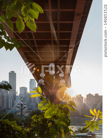 A low-angle, worm's-eye view captures the massive geometric grid underbelly of Chongqing's orange Dongshuimen Bridge framing a distant skyscraper skyline, green foliage, and a multi-globe street lamp A low-angle, worm's-eye view captures the massive geometric grid underbelly of Chongqing's orange Dongshuimen Bridge framing a distant skyscraper skyline, green foliage, and a multi-globe street lamp 140853528