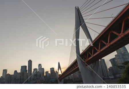 a dramatic low-angle perspective highlights the bold architectural steelwork and sweeping cables of the Qiansimen Bridge as it stretches majestically over the river toward a hazy, high-rise cityscape 140853535