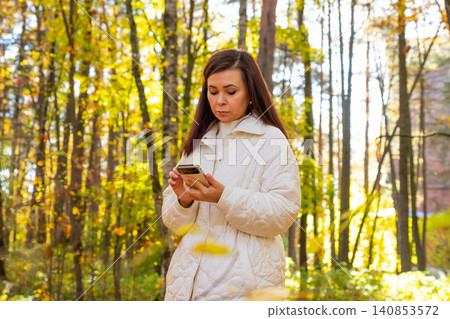 Woman 42 years old looking at smartphone in autumn forest. Caucasian lady using mobile device for digital communication during walking in park. Leisure activity on sunny fall day outdoors. Woman 42 years old looking at smartphone in autumn forest. Caucasian lady using mobile device for digital communication during walking in park. Leisure activity on sunny fall day outdoors. 140853572