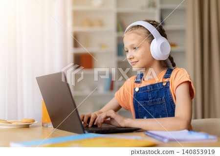Teen girl in headphones typing on laptop at home, studying beside notebooks Teen girl in headphones typing on laptop at home, studying beside notebooks 140853919