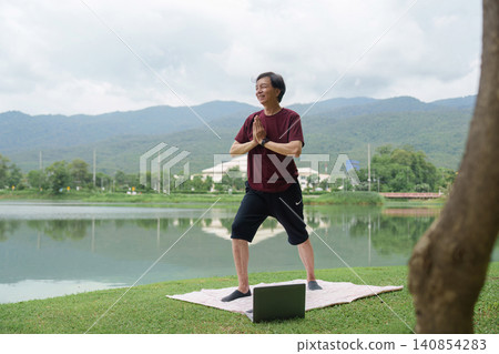 Active senior Asian man practicing yoga outdoors by lake with laptop Active senior Asian man practicing yoga outdoors by lake with laptop 140854283