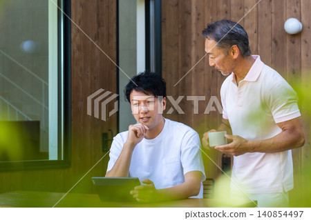 一對年邁的父親和兒子在露台上一邊看著平板電腦一邊討論未來。 一對年邁的父親和兒子在露台上一邊看著平板電腦一邊討論未來。 140854497