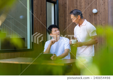 一對年邁的父親和兒子在露台上一邊看著平板電腦一邊討論未來。 一對年邁的父親和兒子在露台上一邊看著平板電腦一邊討論未來。 140854498