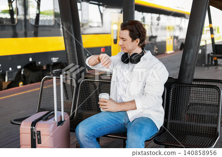 man with a suitcase checking his watch on a railway platform. He awaits eco-friendly rail travel, supporting low-carbon transport and eco-mobility. 140858956