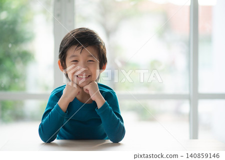 Portrait of a happy smiling Asian little boy sitting at the table and resting chin on hands indoors. Portrait of a happy smiling Asian little boy sitting at the table and resting chin on hands indoors. 140859146