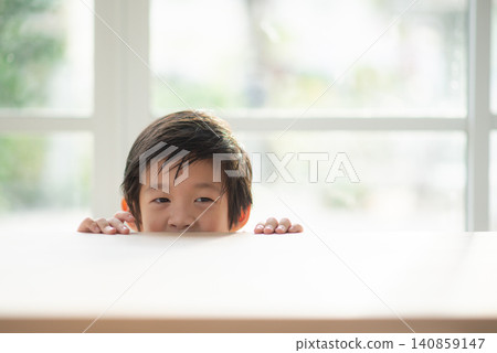 Cute Asian little boy with wet hair peeking over the edge of a wooden table indoors with copy space. Cute Asian little boy with wet hair peeking over the edge of a wooden table indoors with copy space. 140859147