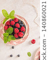 A high angle shot displays a rustic bowl brimming with fresh raspberries and blueberries decorated with mint sprigs on a floral vintage plate over a bright textured surface with scattered fruit 140860021