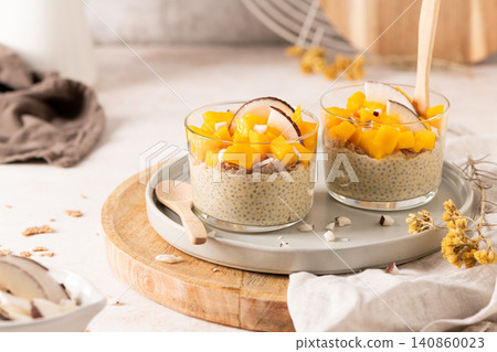 Two glasses of creamy chia seed pudding topped with vibrant diced mango and fresh coconut slices served on a ceramic plate with wooden spoons for a healthy breakfast or nutritious tropical snack Two glasses of creamy chia seed pudding topped with vibrant diced mango and fresh coconut slices served on a ceramic plate with wooden spoons for a healthy breakfast or nutritious tropical snack 140860023