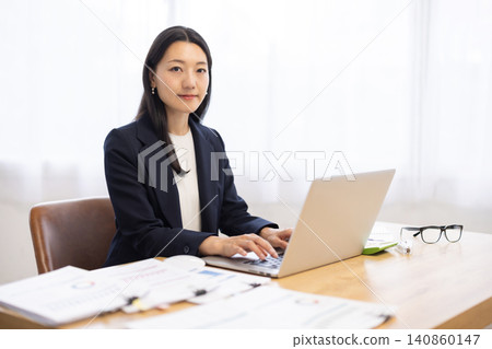 一位日本女商人對著鏡頭微笑，她坐在明亮的窗邊使用筆記型電腦工作。 140860147