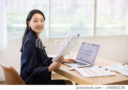 A Japanese businesswoman smiles at the camera while working on her laptop by a bright window. A Japanese businesswoman smiles at the camera while working on her laptop by a bright window. 140860156