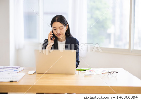 A young Japanese businesswoman is talking on her smartphone while using her laptop in a bright window. A young Japanese businesswoman is talking on her smartphone while using her laptop in a bright window. 140860394