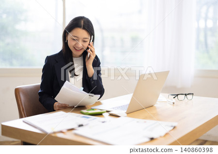 A young Japanese businesswoman is talking on her smartphone while using her laptop in a bright window. 140860398
