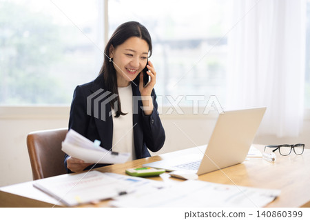A young Japanese businesswoman is talking on her smartphone while using her laptop in a bright window. A young Japanese businesswoman is talking on her smartphone while using her laptop in a bright window. 140860399