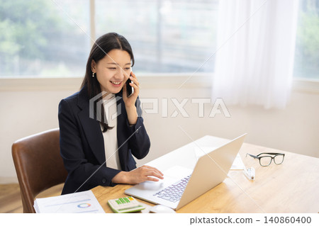 一位年輕的日本女商人正在明亮的窗邊一邊用筆記型電腦一邊用智慧型手機通話。 一位年輕的日本女商人正在明亮的窗邊一邊用筆記型電腦一邊用智慧型手機通話。 140860400