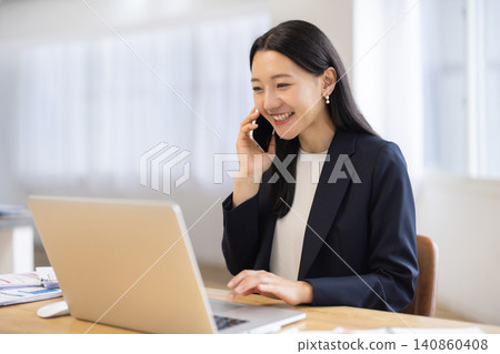 A young Japanese businesswoman is talking on her smartphone while using her laptop in a bright window. A young Japanese businesswoman is talking on her smartphone while using her laptop in a bright window. 140860408