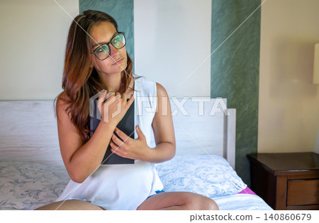 Woman wearing glasses sitting casually on a bed in her bedroom, hugging a book while looking up and thinking, reflecting a moment of quiet contemplation or dreaming Woman wearing glasses sitting casually on a bed in her bedroom, hugging a book while looking up and thinking, reflecting a moment of quiet contemplation or dreaming 140860679