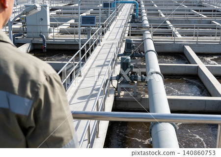Worker overseeing industrial water treatment facility with complex piping and aeration tanks Worker overseeing industrial water treatment facility with complex piping and aeration tanks 140860753