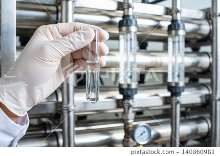 Scientist in lab coat and gloves holds test tube with clear liquid near industrial water filtration system Scientist in lab coat and gloves holds test tube with clear liquid near industrial water filtration system 140860981
