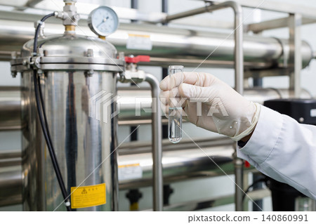 Scientist in lab coat examines liquid in test tube near industrial filtration equipment Scientist in lab coat examines liquid in test tube near industrial filtration equipment 140860991