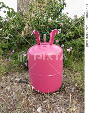 A pink portable gas cylinder tank is placed on dry soil in an outdoor area. It stands in front of a bush with blooming pink flowers. This setup could be used for a party or refrigerant idea. 140861541