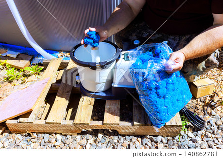 Workman holds bag of blue filter material while setting up pool cleaning machine in an outdoor area. 140862781