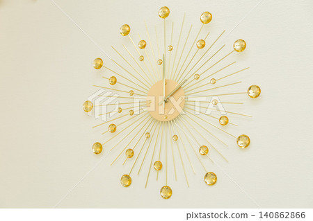 A gold sunburst clock hanging on a white wall, mid-century style. A gold sunburst clock hanging on a white wall, mid-century style. 140862866