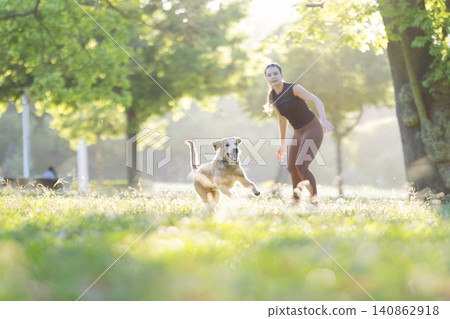 Woman in athletic wear running alongside a golden retriever dog in a sunlit park with trees and grass, showcasing an active outdoor lifestyle 140862918