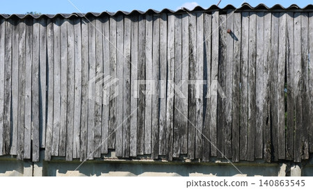 An old wooden fence wall of gray vertical boards. The faded wood is cracked and grained with age. Above, the corrugated slate roof of a barn is visible, and below, a concrete foundation and greenery. An old wooden fence wall of gray vertical boards. The faded wood is cracked and grained with age. Above, the corrugated slate roof of a barn is visible, and below, a concrete foundation and greenery. 140863545