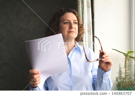 Middle-aged woman reading a document by the window holding eyewear and looking thoughtful Middle-aged woman reading a document by the window holding eyewear and looking thoughtful 140863707