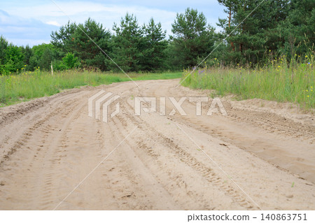 Sandy country road through a field turns into the forest. Dirt road stretches past a pine forest under an overcast sky Sandy country road through a field turns into the forest. Dirt road stretches past a pine forest under an overcast sky 140863751