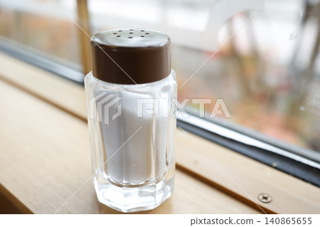 窗邊放著一個鹽瓶,棕色的蓋子,以及一個有孔的調味料容器。 窗邊放著一個鹽瓶,棕色的蓋子,以及一個有孔的調味料容器。 140865655