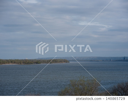 Volga river near Volgograd city. Wide-angle view of a calm coastal wetland featuring an island surrounded by water under a cloudy sky. Volga river near Volgograd city. Wide-angle view of a calm coastal wetland featuring an island surrounded by water under a cloudy sky. 140865729