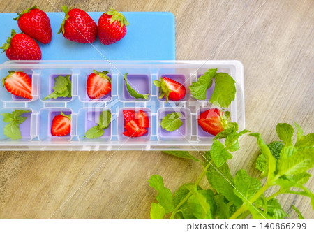 Preparing ice cubes with fresh strawberries and mint leaves in a plastic tray on a wooden table. High angle view. Summer refreshing drinks ingredients. Fresh strawberry and green mint in ice cube tray Preparing ice cubes with fresh strawberries and mint leaves in a plastic tray on a wooden table. High angle view. Summer refreshing drinks ingredients. Fresh strawberry and green mint in ice cube tray 140866299