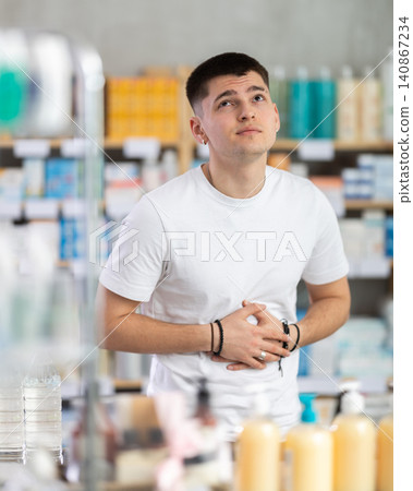 Sick young man buyer with stomach pain choosing medicines and hygiene products in pharmacy. Health care and medicine concept Sick young man buyer with stomach pain choosing medicines and hygiene products in pharmacy. Health care and medicine concept 140867234